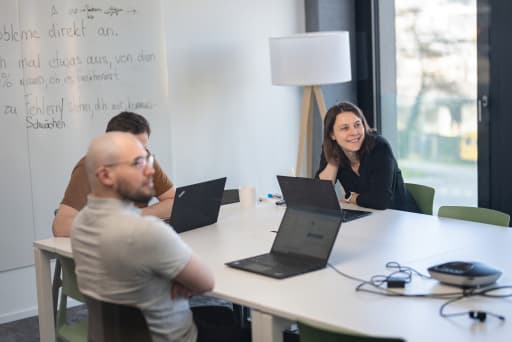 Eine Mitarbeiterin von Xamplo sitzt lächelnd mit Laptop an einem Tisch in einem hellen Büro.