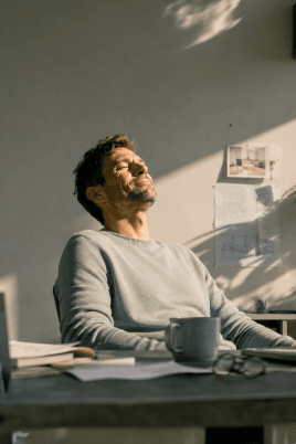 Ein Mann sitzt entspannt an seinem Schreibtisch im Büro, lehnt sich zurück und geniesst einen Moment im Sonnenlicht.