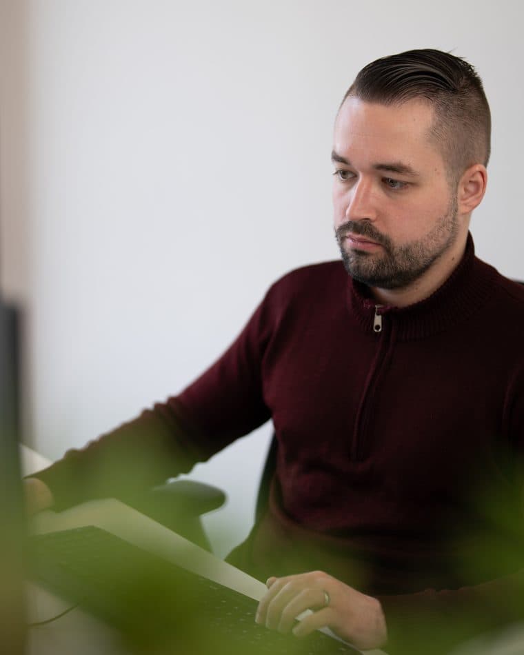 Platzhalter-Teamfoto (Stockfoto): ein Mann in dunkelrotem Pullover arbeitet konzentriert am Bildschirm — echter Name und Foto folgen vor Live-Gang (siehe AGENTS.md Abschnitt 7).