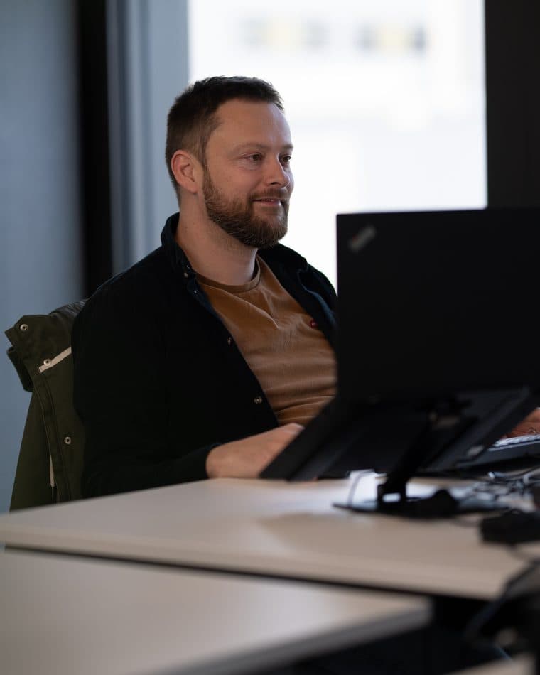 Platzhalter-Teamfoto (Stockfoto): ein Mann in dunkler Jacke sitzt lächelnd vor seinem Laptop im Büro — echter Name und Foto folgen vor Live-Gang (siehe AGENTS.md Abschnitt 7).