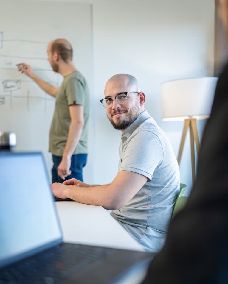 Ein Xamplo-Mitarbeiter sitzt lächelnd am Laptop, im Hintergrund beschriftet ein Kollege ein Whiteboard mit einer Prozessskizze.